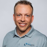 A person wearing a light gray polo shirt with a logo on the left side, posing against a plain white background.